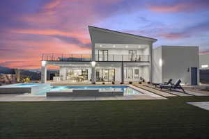 Back house at dusk featuring a patio, a balcony, a lawn, and a pool with hot tub