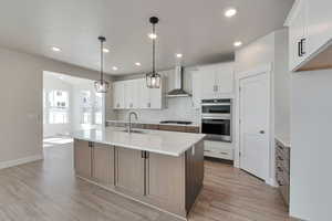 Kitchen featuring sink, white cabinetry, stainless steel double oven, a kitchen island with sink, and wall chimney range hood