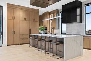 Kitchen with a breakfast bar, light countertops, wall chimney exhaust hood, light wood finished floors, and a center island with sink