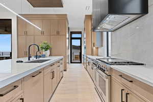 Kitchen with range with two ovens, light stone counters, light brown cabinets, a sink, and wall chimney range hood