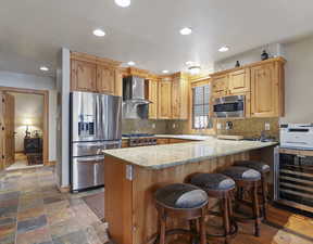 Kitchen with wine cooler, light stone counters, light brown cabinets, appliances with stainless steel finishes, and wall chimney range hood