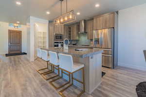 Kitchen featuring stainless steel appliances, a center island with sink, decorative light fixtures, wall chimney exhaust hood, and light wood-type flooring