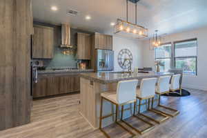 Kitchen with pendant lighting, an island with sink, sink, stainless steel fridge with ice dispenser, and wall chimney range hood