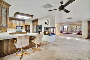 Kitchen with tile countertops, light carpet, stainless steel refrigerator, a multi sided fireplace, and a kitchen breakfast bar