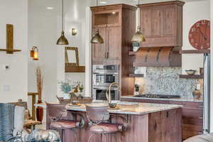 Kitchen with light stone counters, backsplash, open shelves, a kitchen island with sink, and appliances with stainless steel finishes