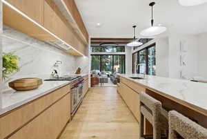 Kitchen with light stone countertops, modern cabinets, light brown cabinets, double oven range, and decorative light fixtures
