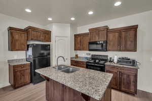 Kitchen with an island with sink, light wood-type flooring, black appliances, light stone counters, and sink