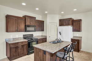 Kitchen featuring light stone countertops, black appliances, sink, a kitchen island with sink, and dark brown cabinets