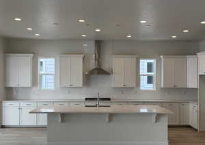 Kitchen with a kitchen island with sink, wall chimney range hood, a healthy amount of sunlight, and white cabinets