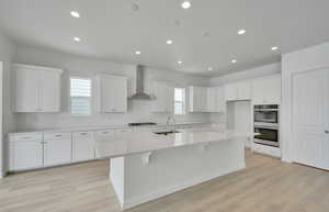 Kitchen featuring sink, white cabinetry, wall chimney exhaust hood, a center island with sink, and stainless steel double oven