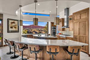 Kitchen featuring a large island, wall chimney range hood, appliances with stainless steel finishes, hanging light fixtures, and light stone counters