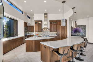 Kitchen with wall chimney range hood, hanging light fixtures, a spacious island, tasteful backsplash, and light stone countertops