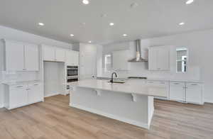 Kitchen with sink, white cabinets, a kitchen island with sink, stainless steel double oven, and wall chimney exhaust hood