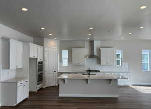 Kitchen with white cabinetry, a wealth of natural light, a center island with sink, and wall chimney range hood