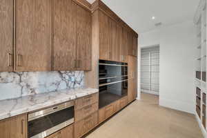 Kitchen featuring stainless steel microwave, decorative backsplash, light stone counters, brown cabinets, and beverage cooler