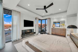 Bedroom with a tray ceiling, access to exterior, ceiling fan, a glass covered fireplace, and light wood-type flooring