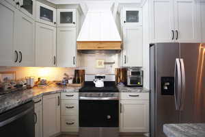 Kitchen with tasteful backsplash, stone countertops, stainless steel appliances, white cabinets, and custom exhaust hood