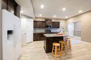 Kitchen featuring appliances with stainless steel finishes, a kitchen bar, light stone counters, dark brown cabinetry, and a center island with sink