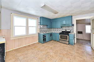 Kitchen with blue cabinetry, stainless steel range with gas cooktop, wooden walls, and decorative backsplash