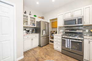 Kitchen with appliances with stainless steel finishes, white cabinets, decorative backsplash, light stone countertops, and light wood-type flooring