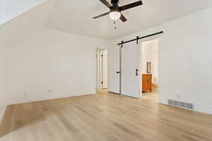 Unfurnished bedroom featuring ceiling fan, ensuite bathroom, a barn door, and light wood-type flooring