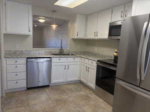 Kitchen with appliances with stainless steel finishes, sink, white cabinets, hanging light fixtures, and light stone counters