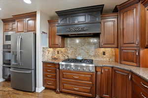 Kitchen with appliances with stainless steel finishes, stone countertops, backsplash, hardwood / wood-style flooring, and custom range hood