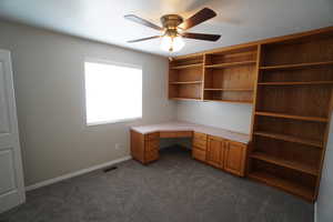 Unfurnished office with dark colored carpet, built in desk, and ceiling fan