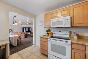 Kitchen with light colored carpet, white appliances, a textured ceiling, light brown cabinetry, and pendant lighting in the dining area