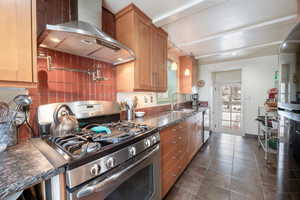 Kitchen featuring sink, dark stone countertops, dark tile patterned flooring, stainless steel appliances, and wall chimney range hood