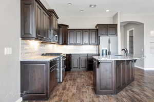 Kitchen with sink, dark wood-type flooring, appliances with stainless steel finishes, dark brown cabinetry, and a center island with sink