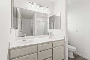 Bathroom featuring toilet, double vanity, a shower with shower door, wood finished floors, and baseboards