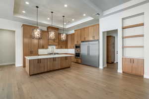 Kitchen featuring built in appliances, pendant lighting, a raised ceiling, light wood-type flooring, and a large island with sink