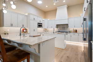 Kitchen featuring white cabinetry, a kitchen island with sink, stainless steel appliances, decorative light fixtures, and light stone counters