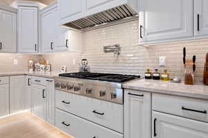 Kitchen with custom exhaust hood, light stone counters, white cabinets, stainless steel gas cooktop, and tasteful backsplash