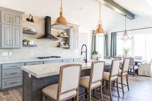 Kitchen with light stone counters, a kitchen island with sink, wall chimney range hood, and pendant lighting