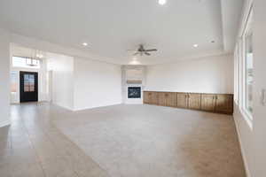 Unfurnished living room with a raised ceiling, light colored carpet, a stone fireplace, and ceiling fan with notable chandelier