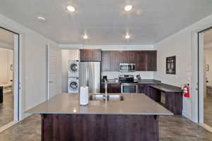 Kitchen featuring sink, stacked washer and clothes dryer, stainless steel appliances, an island with sink, and dark brown cabinets