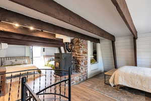 Bedroom featuring beamed ceiling, a wall mounted air conditioner, light hardwood / wood-style flooring, and a wood stove