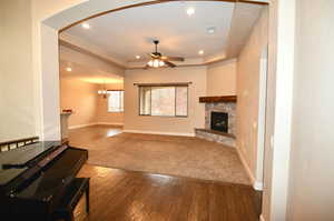 Living room with a tray ceiling, a stone fireplace, wood-type flooring, and ceiling fan with notable chandelier