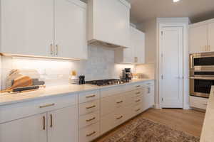 Kitchen featuring stainless steel appliances, white cabinetry, light stone countertops, light hardwood / wood-style floors, and custom exhaust hood