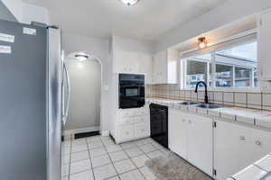 Kitchen featuring tasteful backsplash, tile countertops, white cabinets, black appliances, and sink