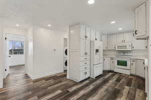 Kitchen with white appliances, stacked washer / dryer, white cabinetry, a textured ceiling, and dark wood-type flooring