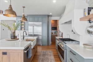 Kitchen with a center island with sink, built in appliances, light stone counters, wood-type flooring, and pendant lighting