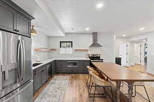 Kitchen with light wood-type flooring, gray cabinetry, a breakfast bar, appliances with stainless steel finishes, and wall chimney exhaust hood