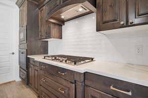 Kitchen featuring stainless steel appliances, backsplash, dark brown cabinets, light stone counters, and custom exhaust hood