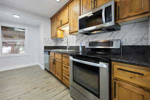 Kitchen featuring light wood-type flooring, dark stone countertops, sink, backsplash, and appliances with stainless steel finishes