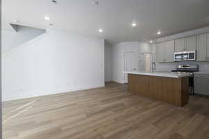 Kitchen with light wood finished floors, a kitchen island with sink, appliances with stainless steel finishes, and gray cabinetry