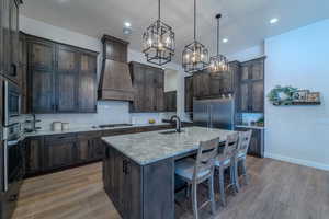 Kitchen with a center island with sink, built in appliances, light stone countertops, premium range hood, and sink