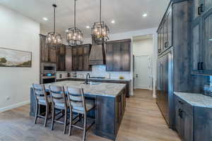 Kitchen featuring an island with sink, custom exhaust hood, dark brown cabinetry, light stone countertops, and appliances with stainless steel finishes